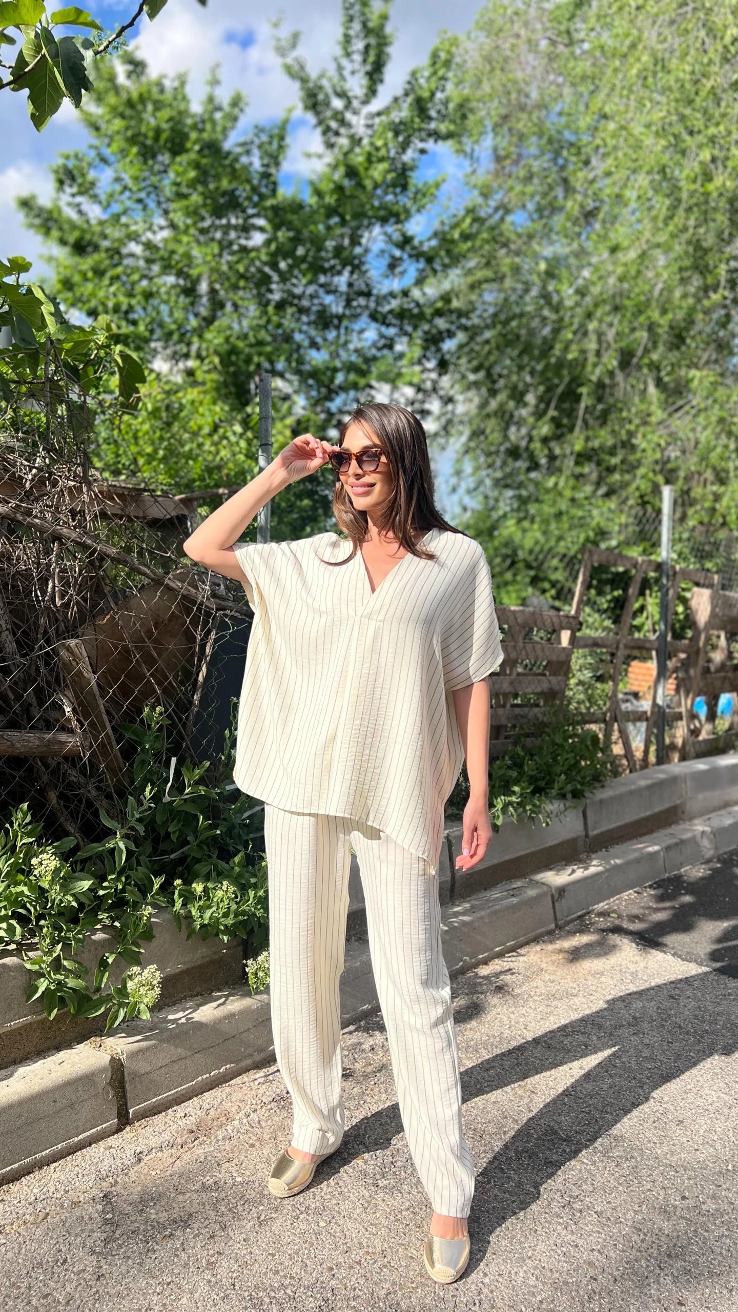 Woman in a white outfit standing outdoors with trees and a fence in the background