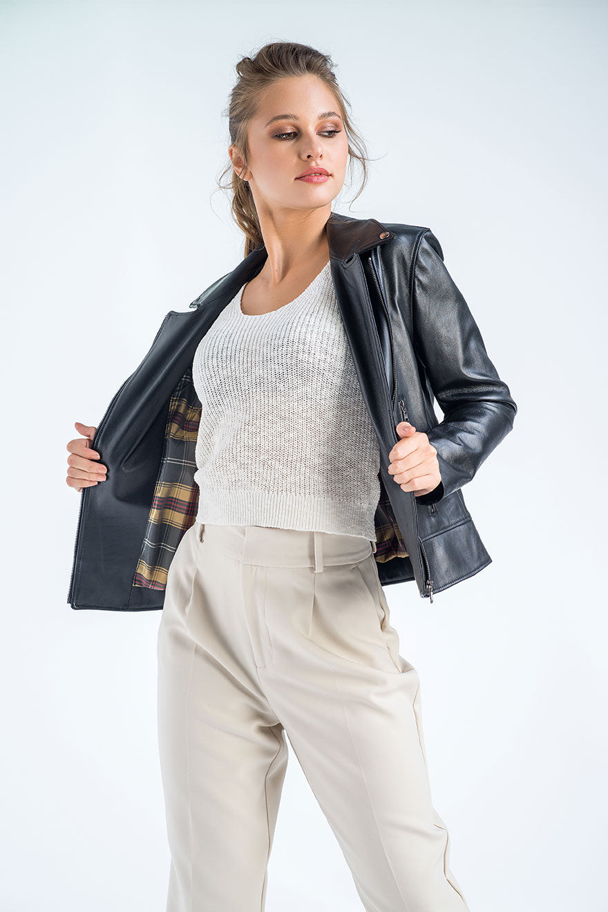 Woman wearing a black leather jacket over a white sweater and beige pants on a light gray background