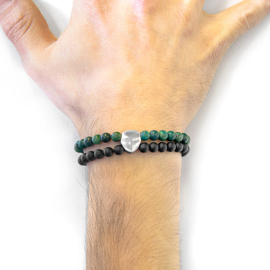 Hand wearing a bracelet with green and black beads and a silver heart charm on a white background