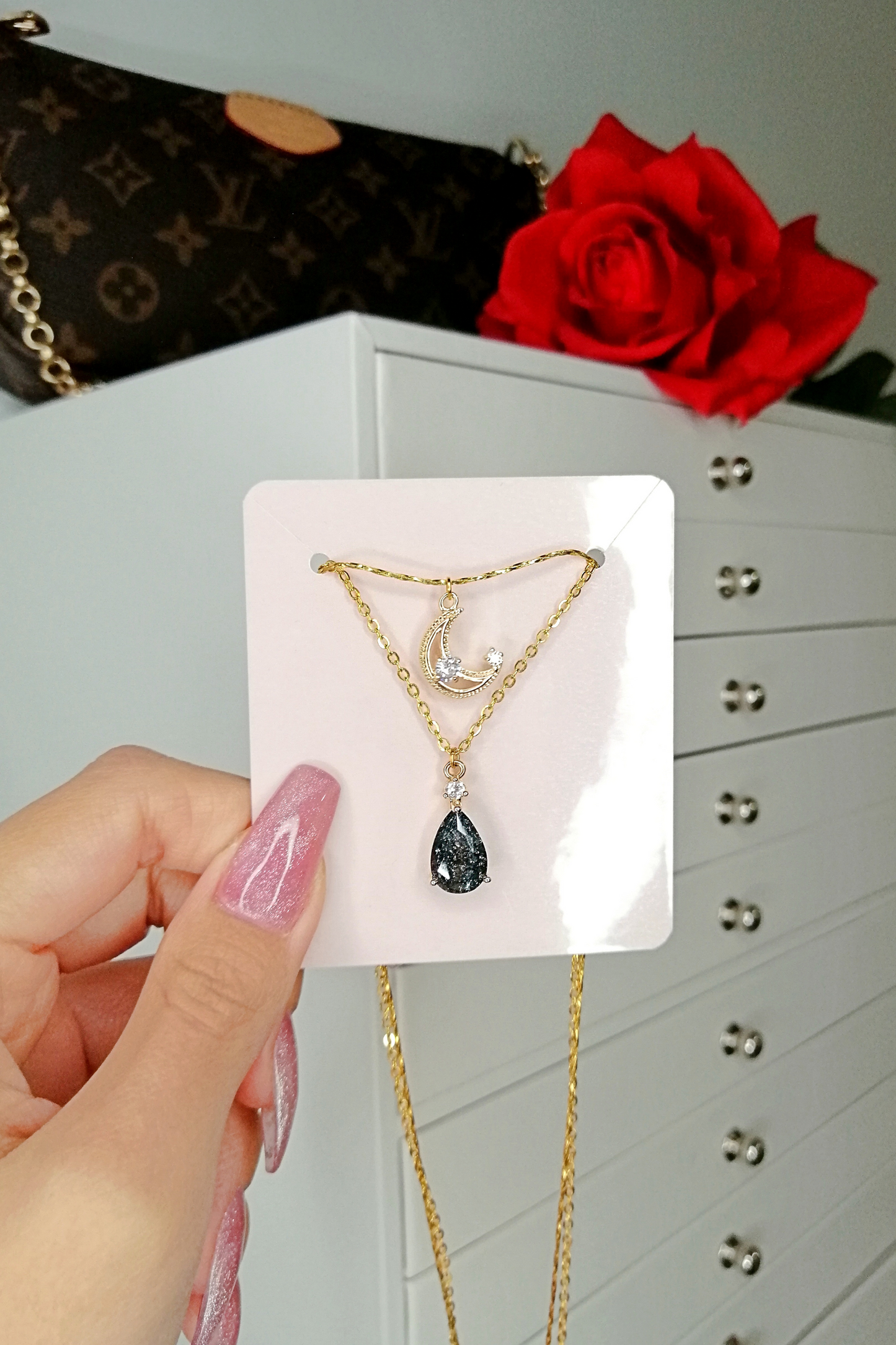 Gold necklace with a black teardrop pendant held by a hand against a neutral background