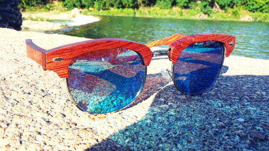Wooden-framed sunglasses with blue reflective lenses on a stone surface near water.