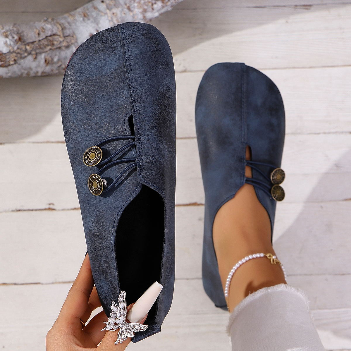 Navy blue flat shoes with gold buttons held by a hand on a light wooden floor.