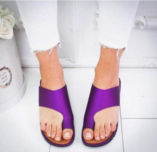 Purple sandals worn with white pants on a white floor.