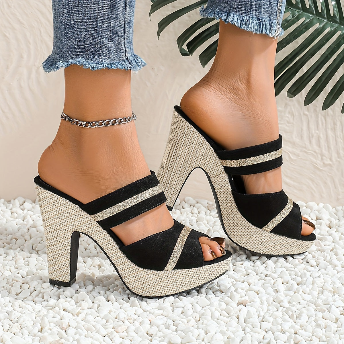 Black and beige strappy high-heeled sandals worn with denim jeans on a textured surface.