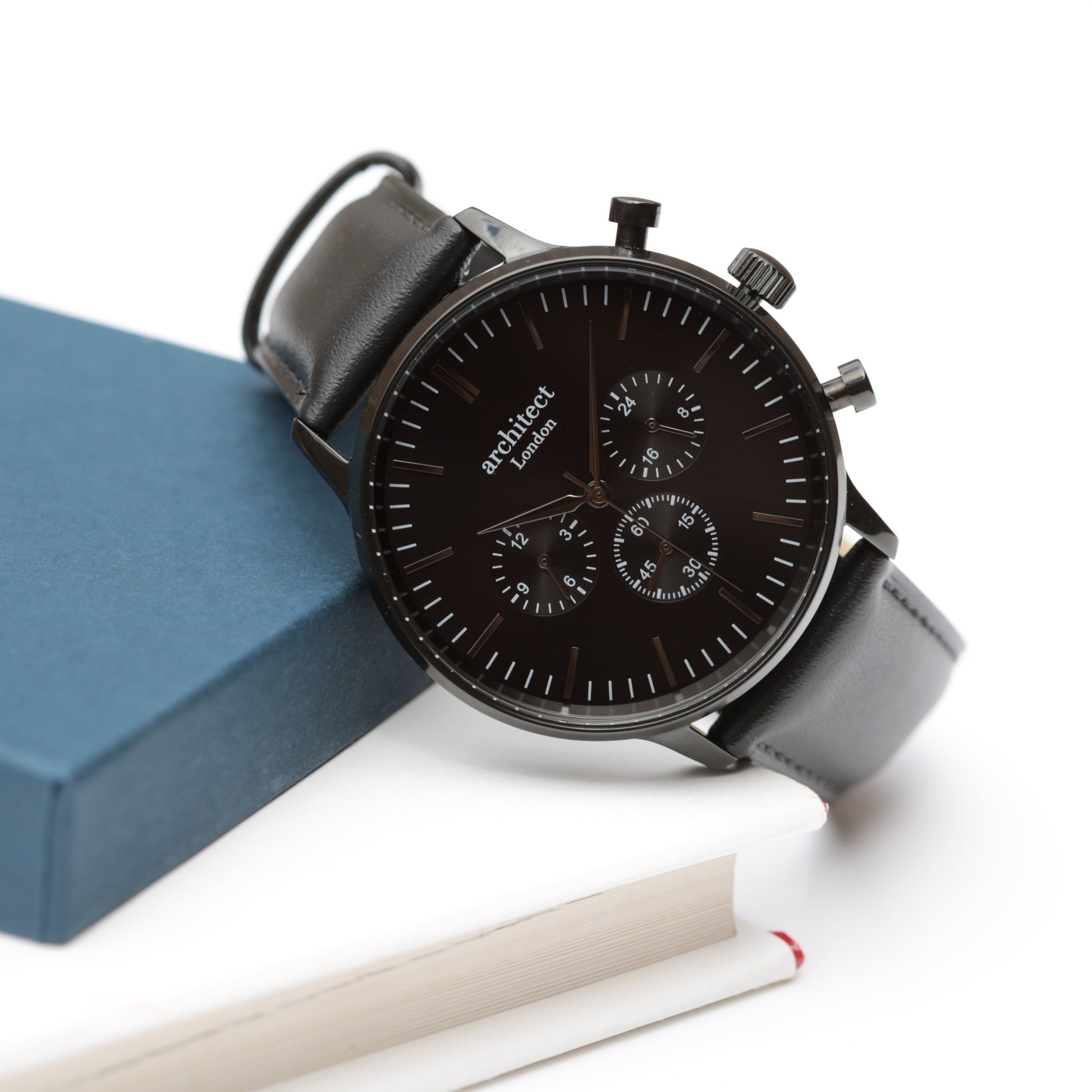 Black watch with leather strap on a white surface with a blue box and white pen.