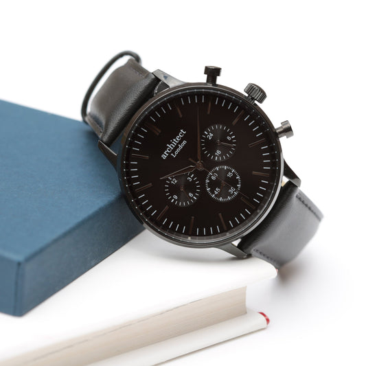 Black watch with leather strap on a white surface with a blue box and white pen.