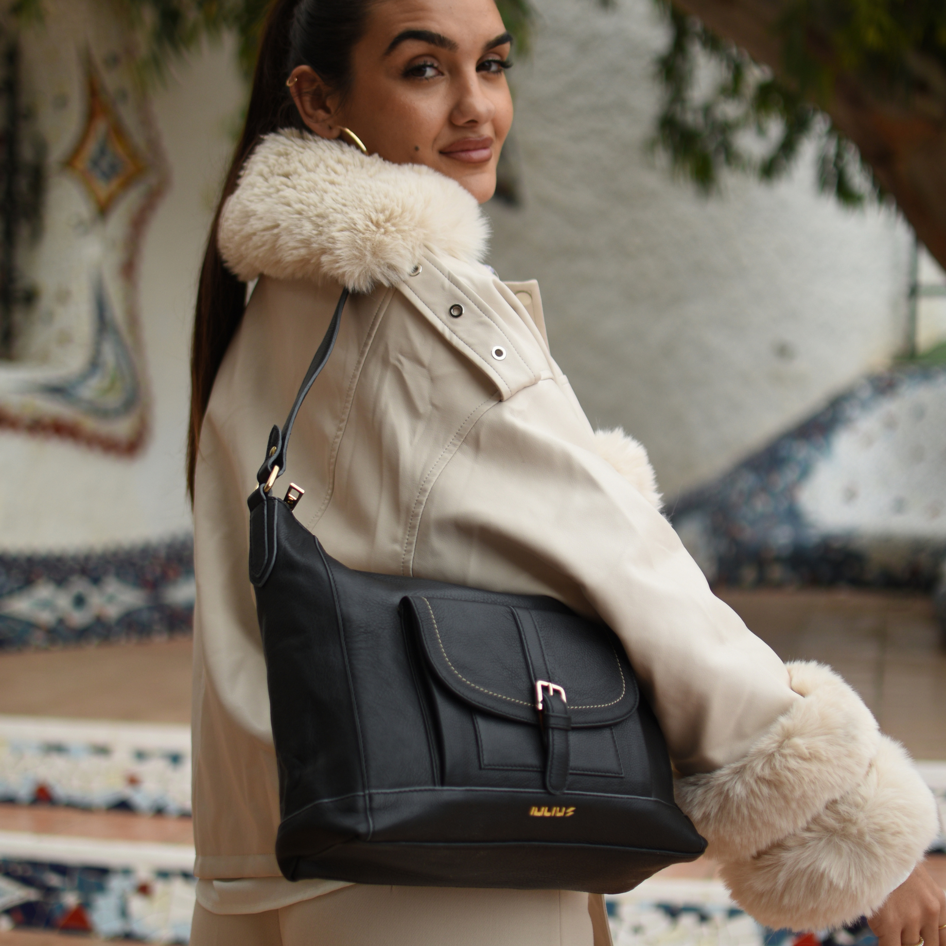Woman wearing a beige coat with fur collar holding a black handbag.