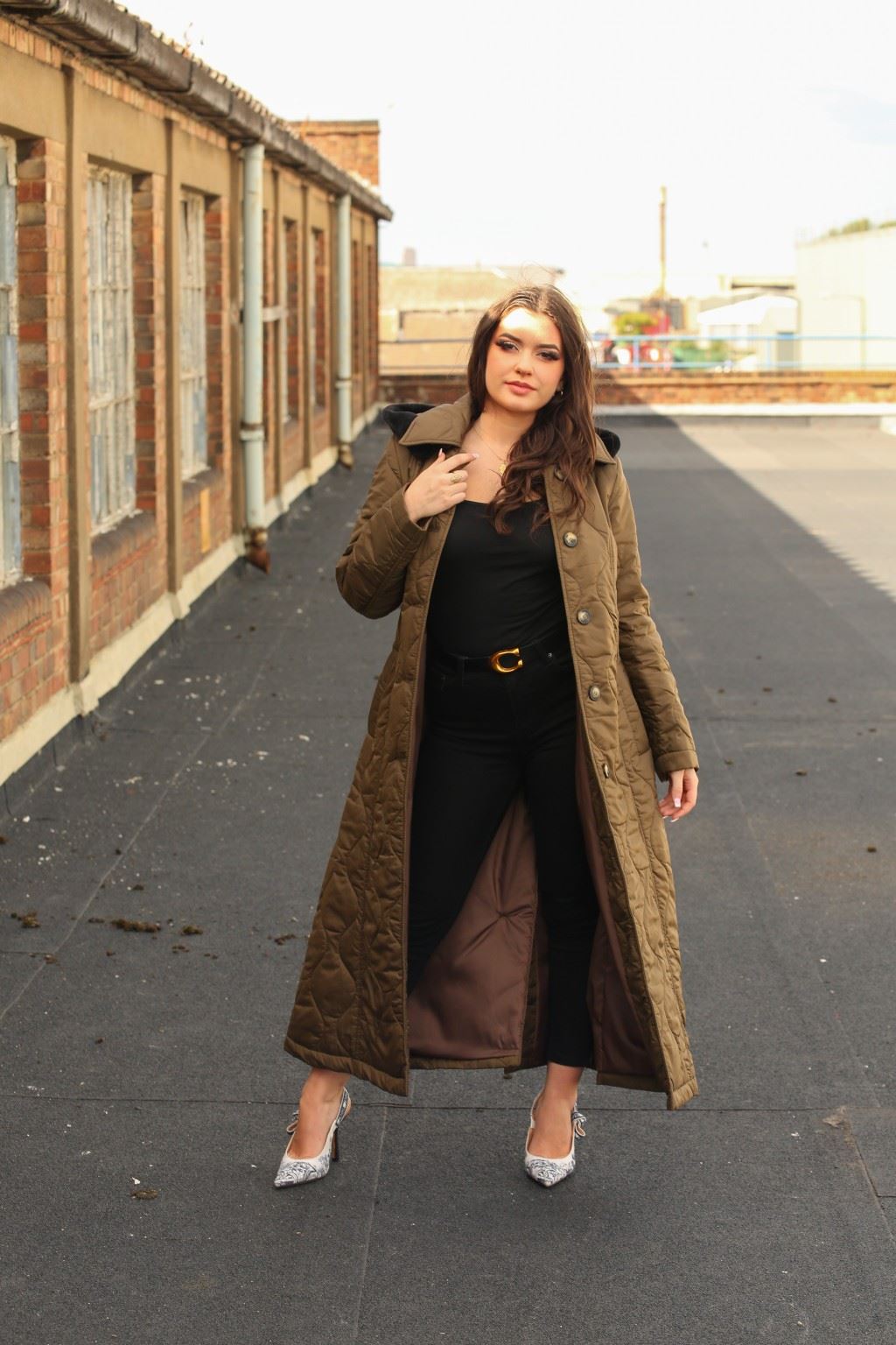 Woman wearing a long brown coat on a rooftop with a building in the background