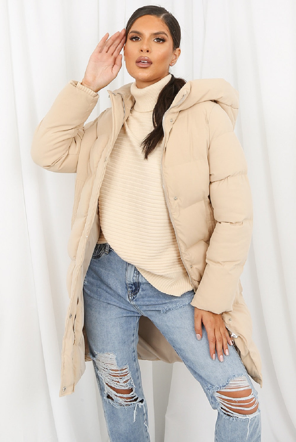 Woman wearing a beige puffer jacket over a beige sweater and blue jeans on a white background