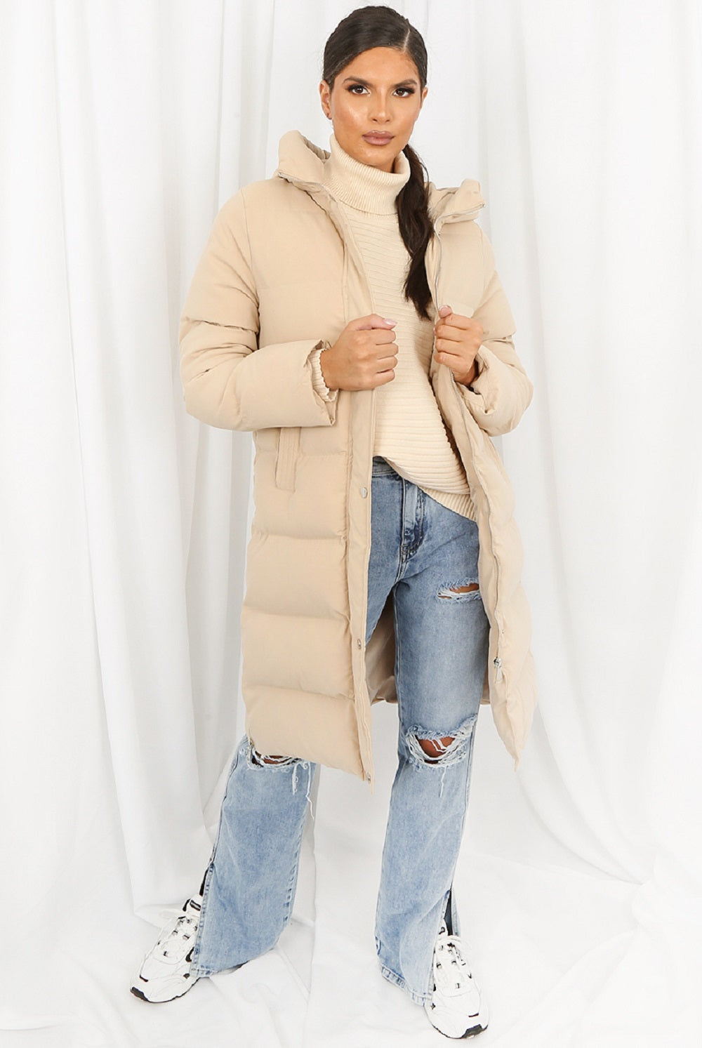Woman wearing a beige puffer coat, white turtleneck, and ripped jeans on a white background