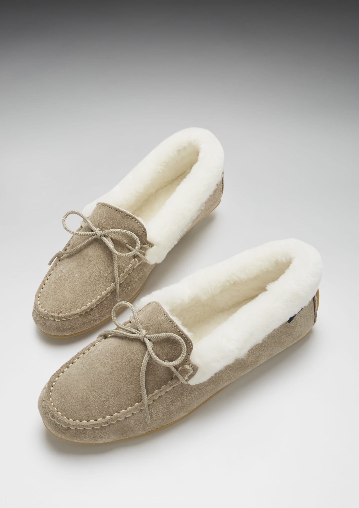 Pair of beige moccasin slippers with white fur lining on a light gray background