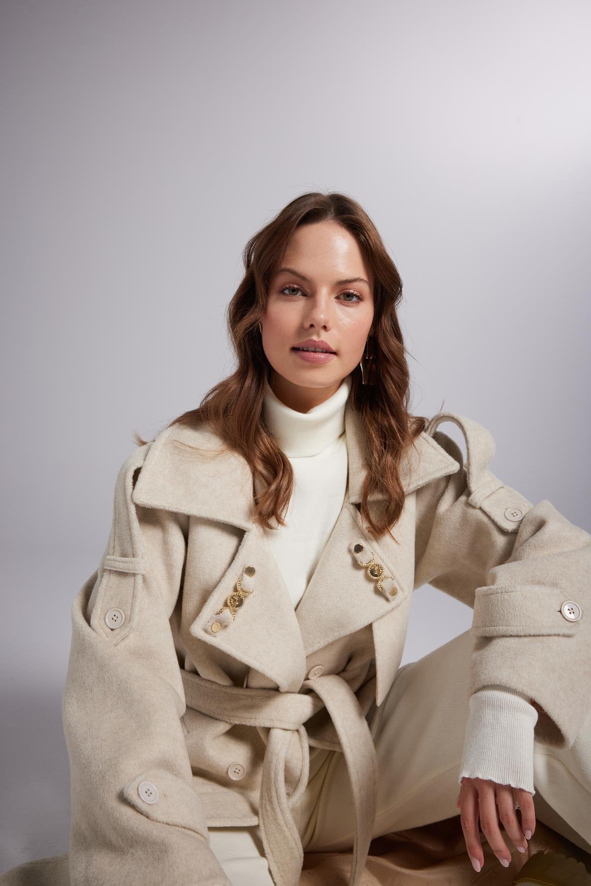 Woman wearing a beige trench coat over a white outfit against a plain background