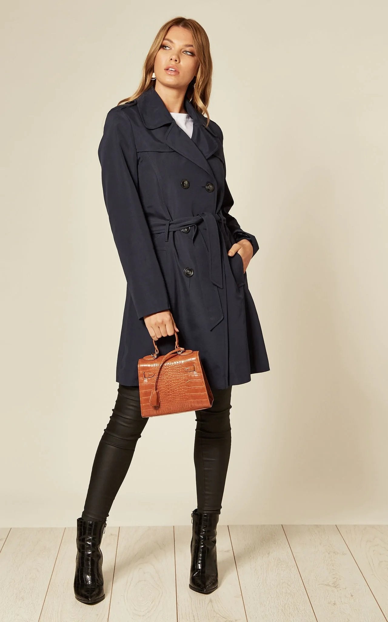 Woman wearing a navy trench coat holding a brown leather bag against a plain background
