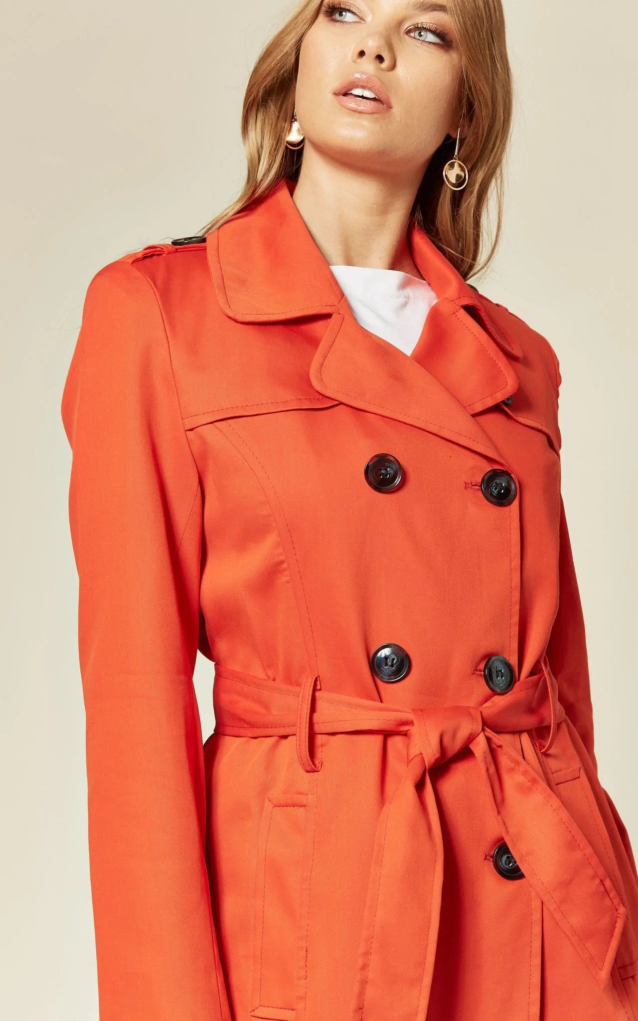 Woman wearing an orange trench coat with black buttons on a beige background