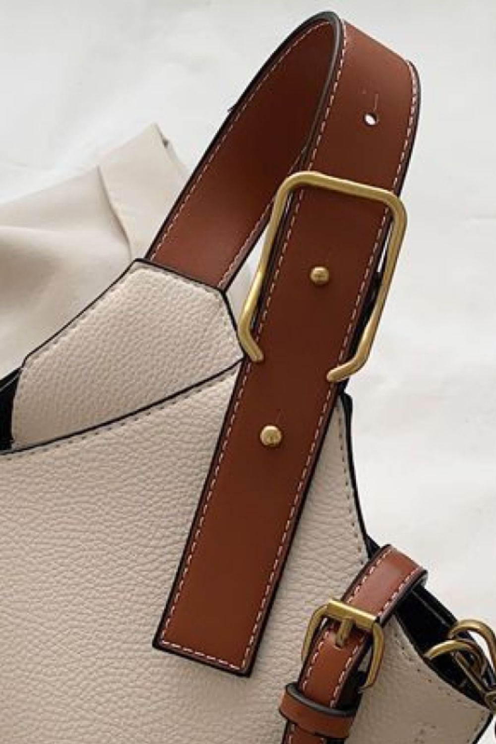 Close-up of a beige handbag with brown leather strap and gold buckle on a light background