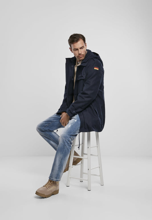 Man sitting on a stool wearing a navy jacket with a brand logo, light blue jeans, and brown boots on a white background.