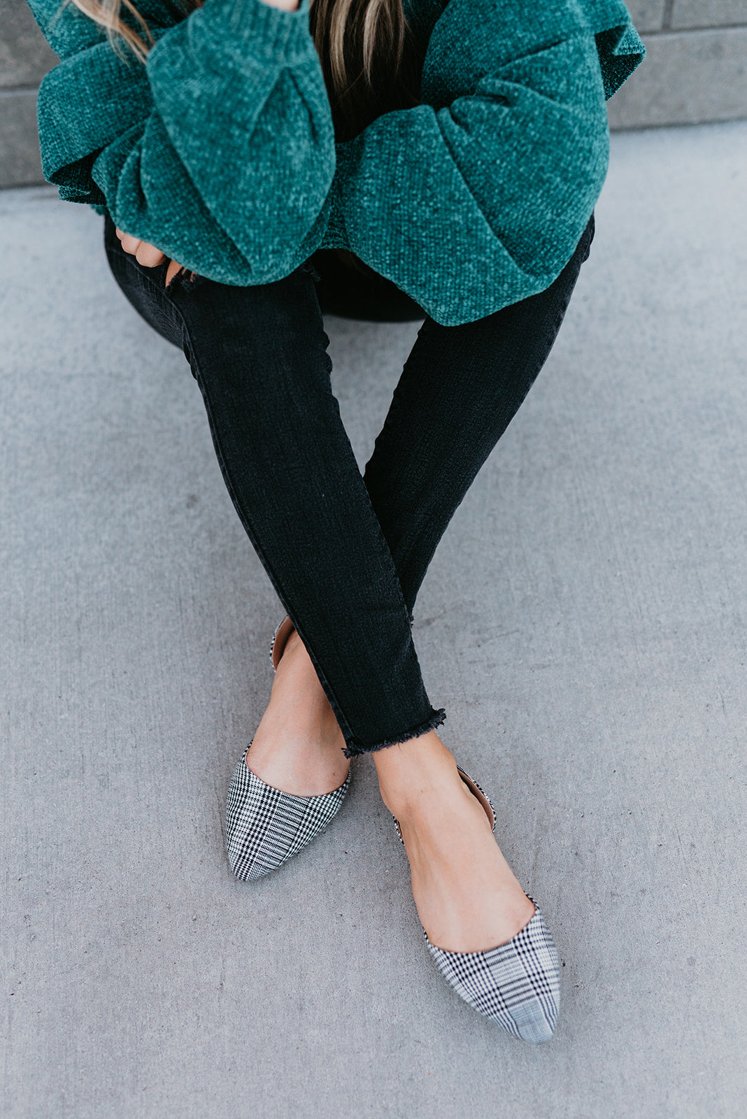 Person wearing a teal sweater, black pants, and plaid shoes on a gray pavement.