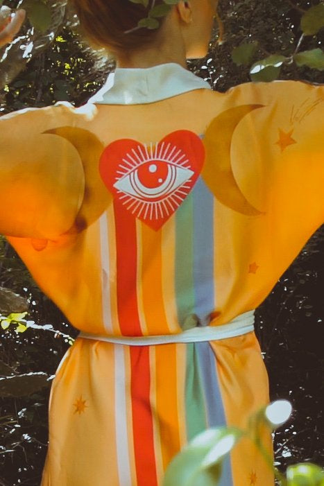 Person wearing a colorful dress with a heart design featuring an eye, surrounded by nature.