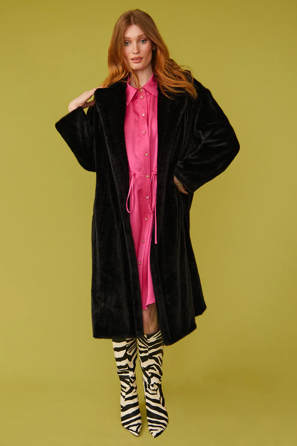 Woman wearing a black coat over a pink dress with zebra-striped boots on a green background