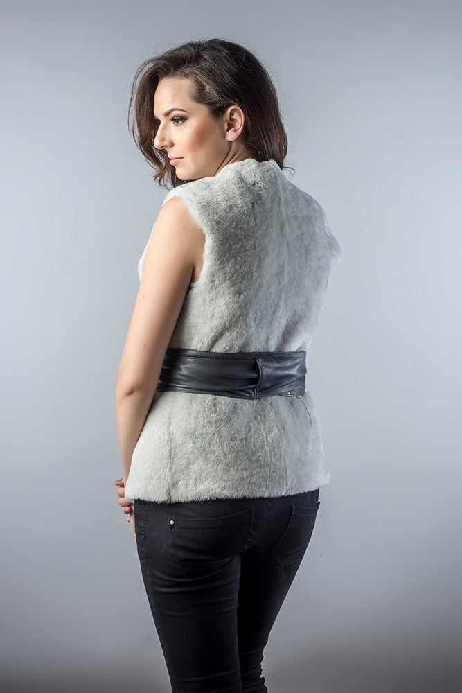 Woman wearing a light gray sleeveless fur vest with a black belt on a plain background