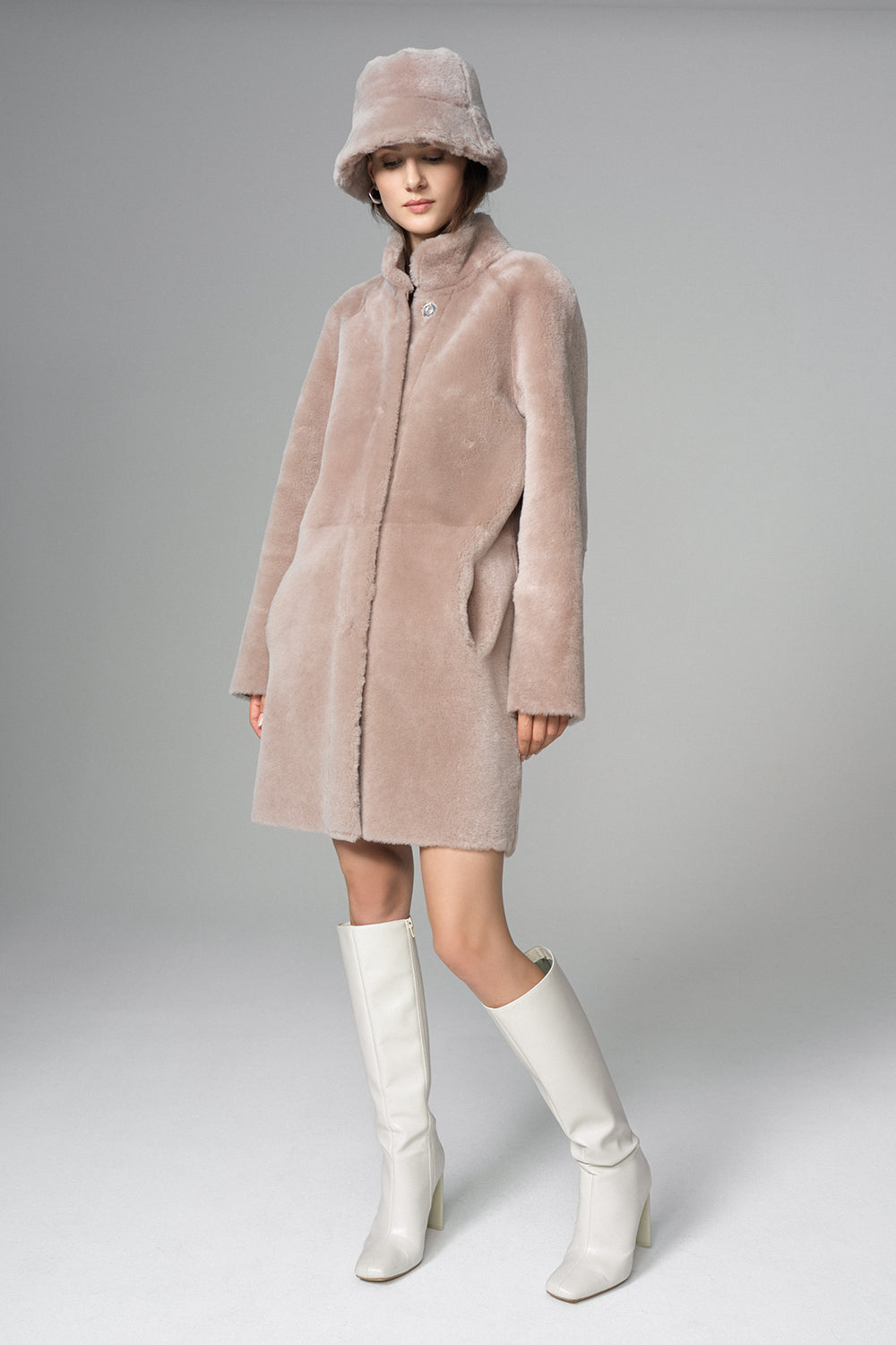 Person wearing a pink fur coat, hat, and white boots on a gray background