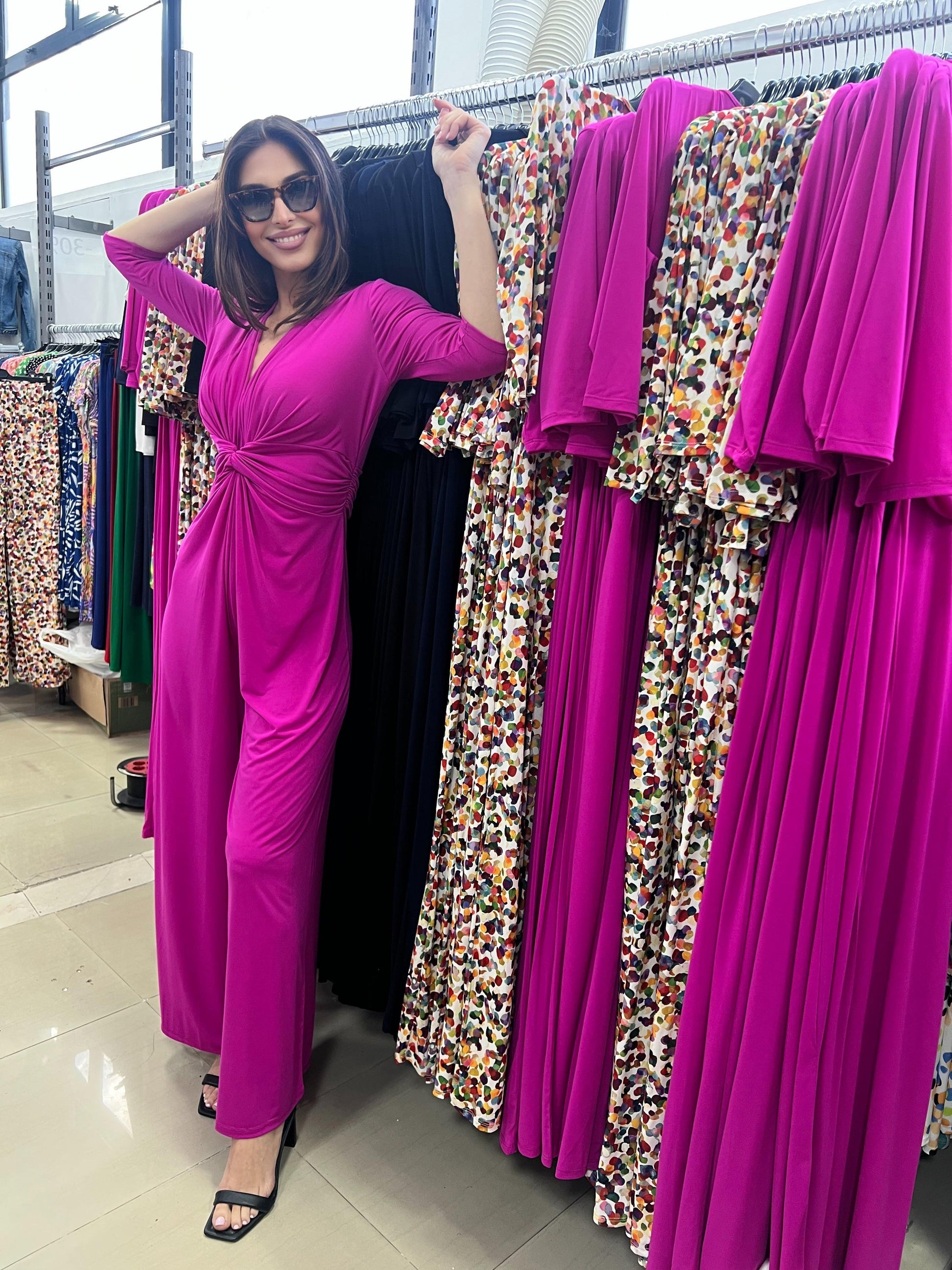 Woman in a bright pink dress standing next to a rack of colorful dresses in a store.