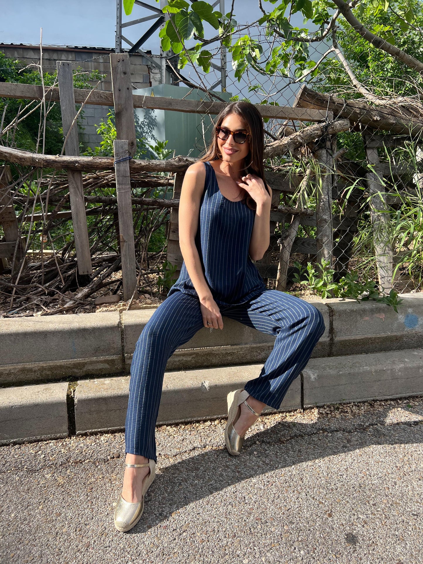 Woman sitting on a concrete ledge wearing a blue outfit with sunglasses, surrounded by greenery.