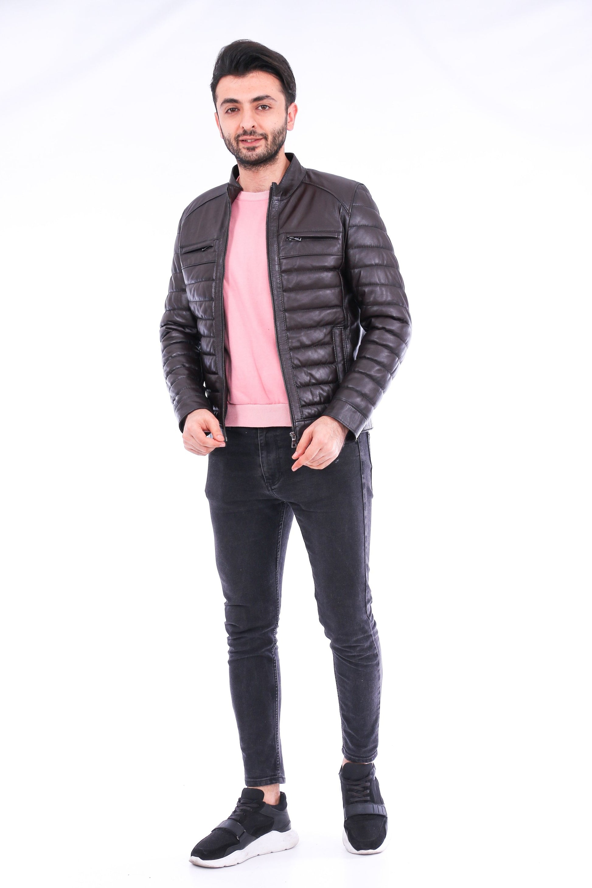 Man wearing a dark gray quilted jacket, pink shirt, black jeans, and black shoes on a white background