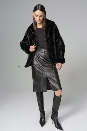 Woman wearing a black fur coat, black top, leather skirt, and black knee-high boots on a gray background