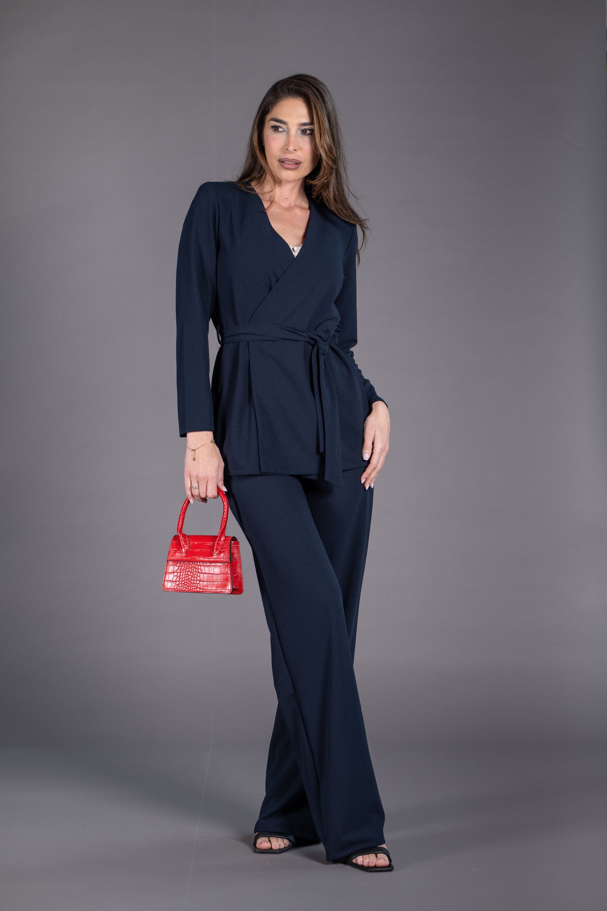 Woman in a navy blue outfit holding a red handbag against a gray background