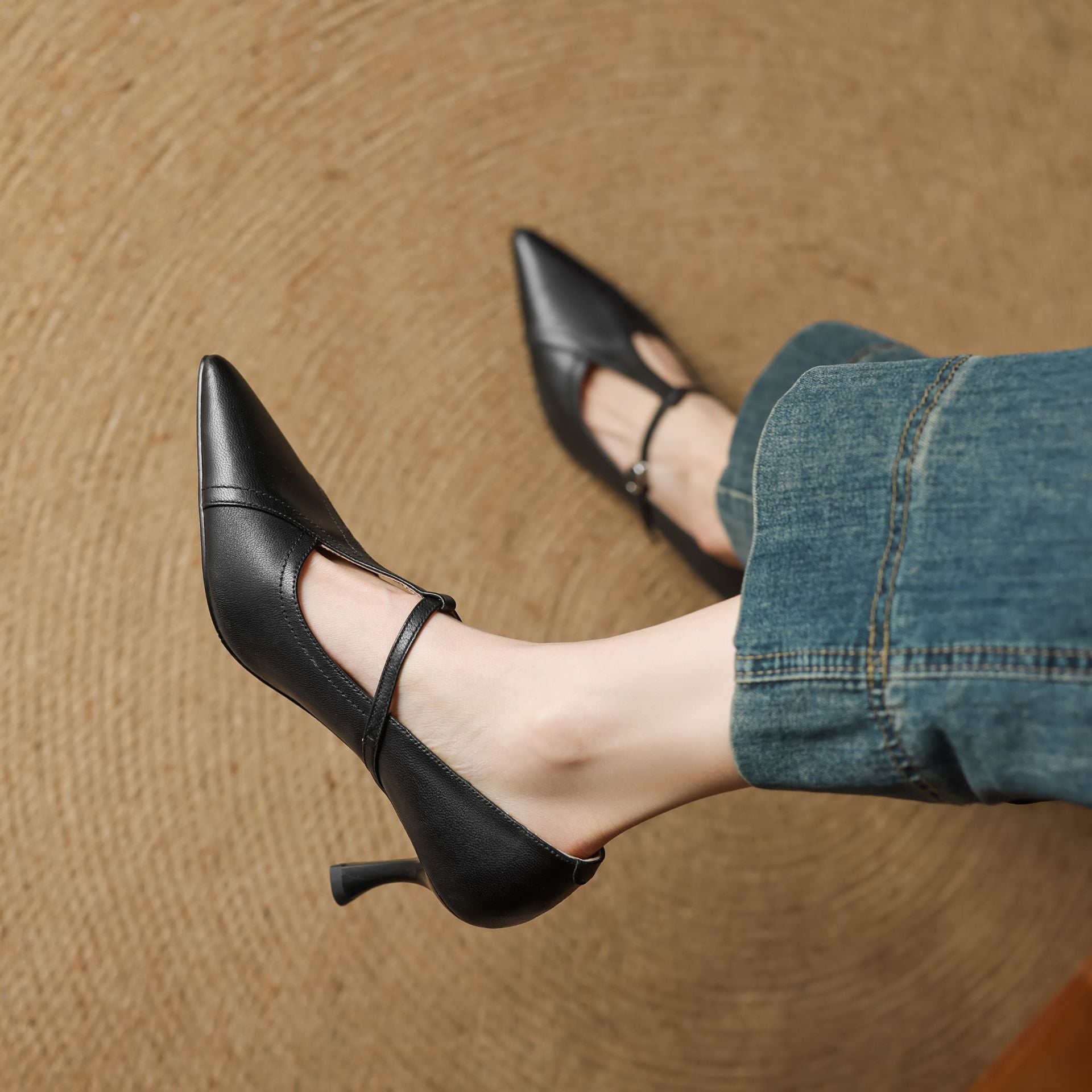 Black high-heeled shoes with thin straps worn with blue jeans on a brown surface