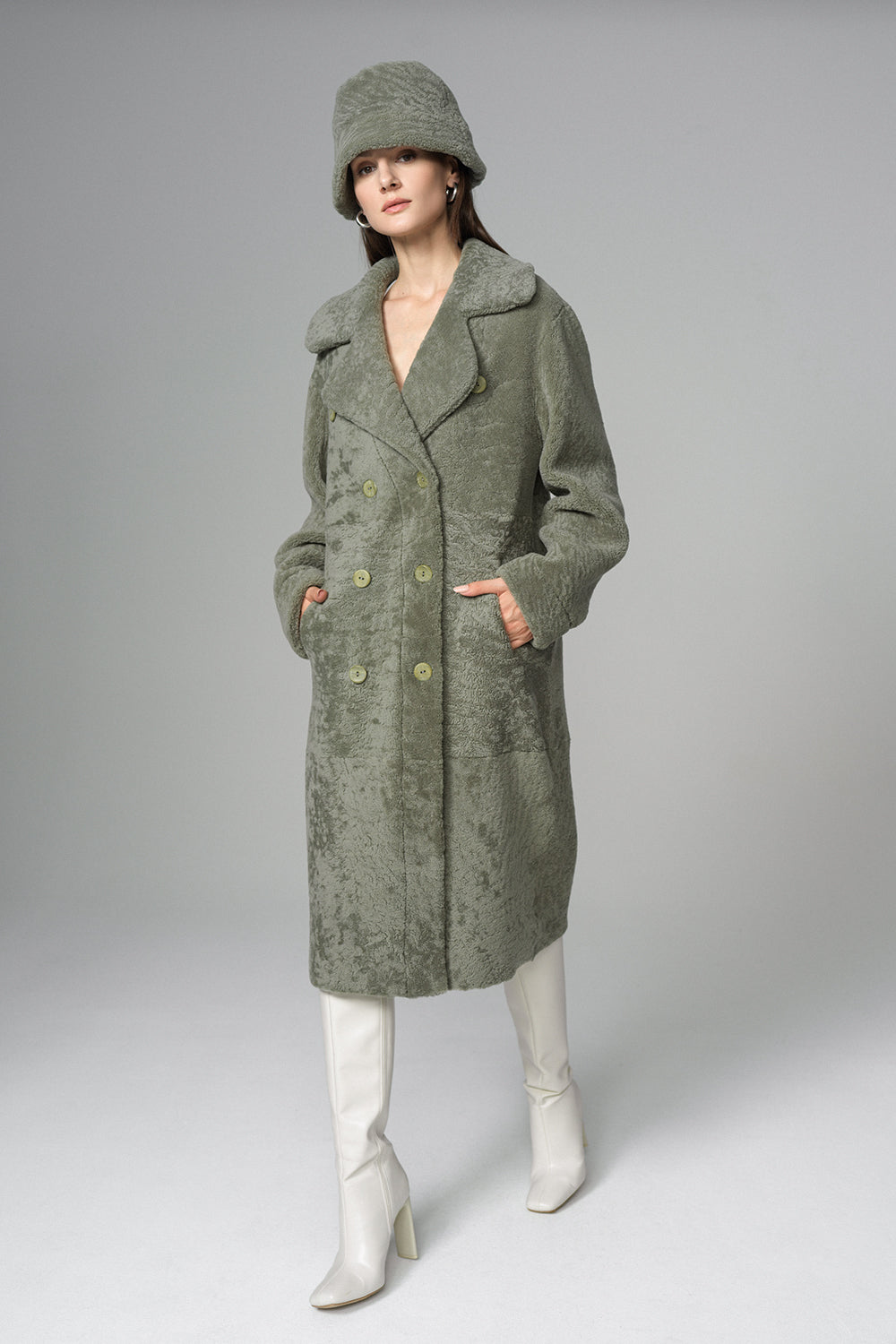 Woman wearing a long green coat, white boots, and a matching hat on a gray background