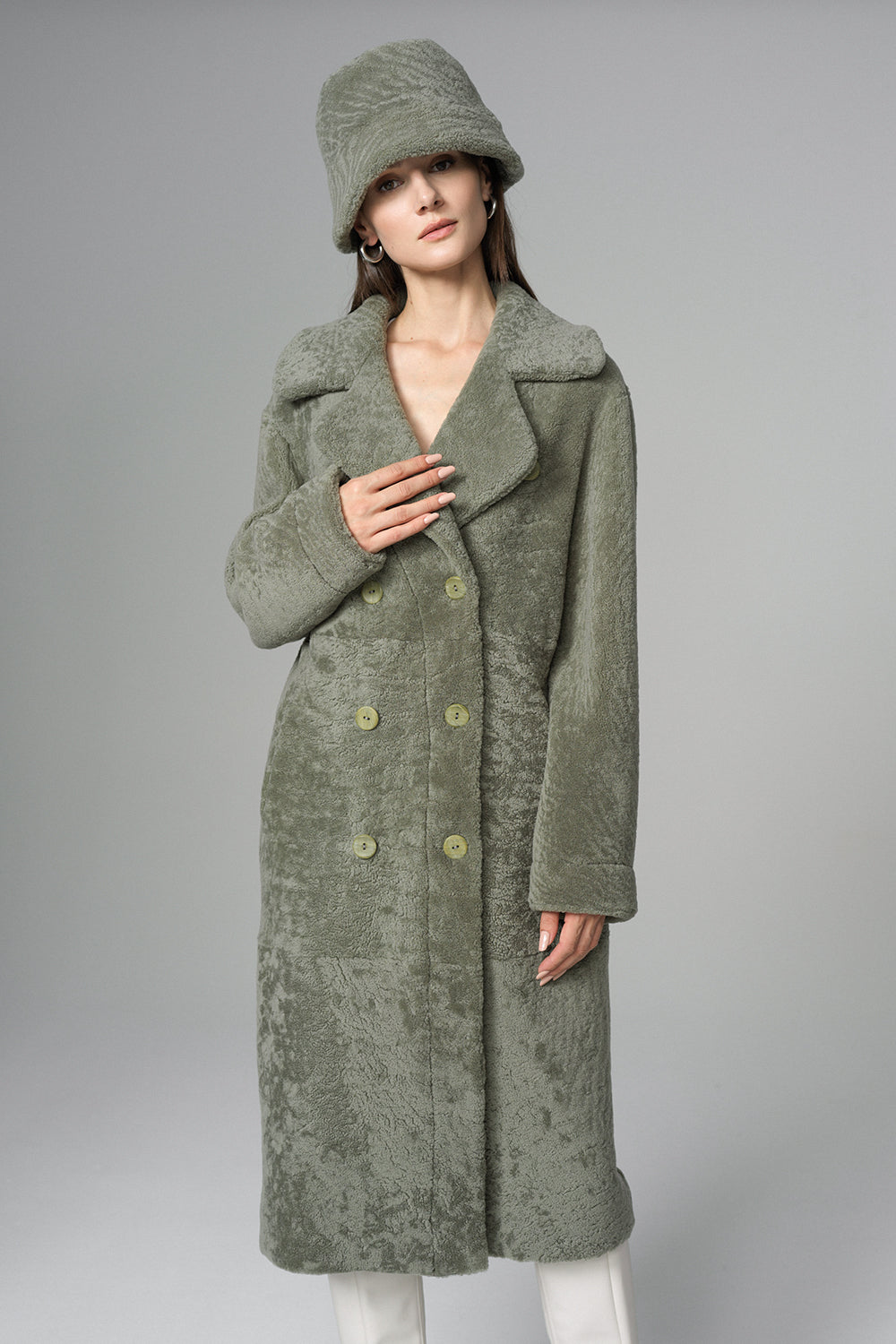 Woman wearing a long green coat and matching hat on a gray background