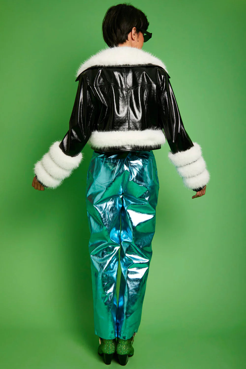 Person wearing a black leather jacket with white fur trim and teal metallic pants on a green background