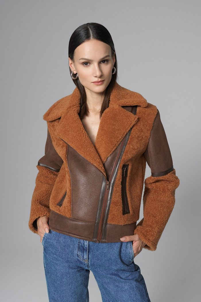 Woman wearing a brown shearling jacket with leather accents on a gray background