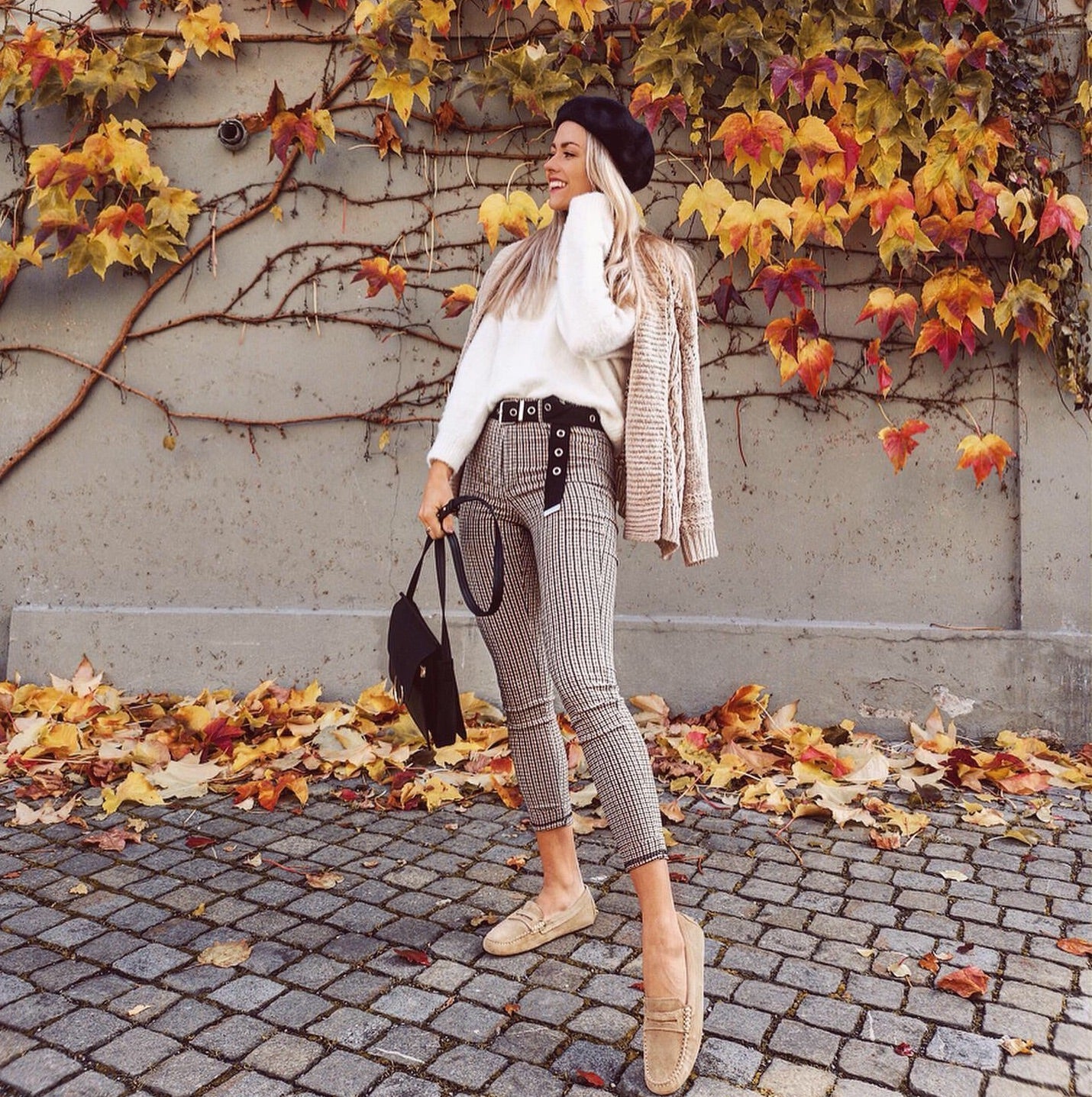Woman in stylish outfit with plaid pants and beige cardigan standing against a wall with autumn leaves.