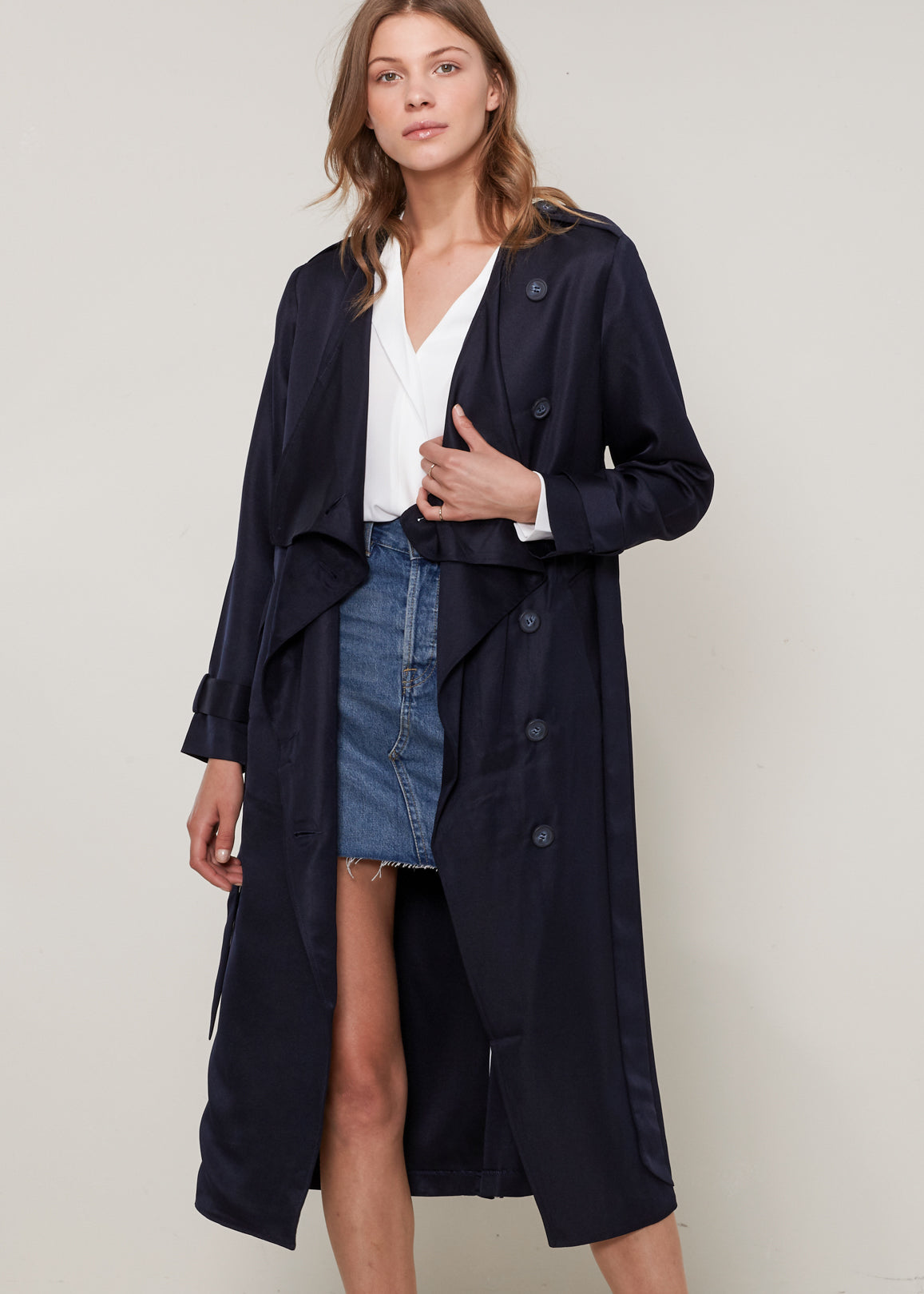 Woman wearing a navy trench coat over a white shirt and denim skirt on a plain background