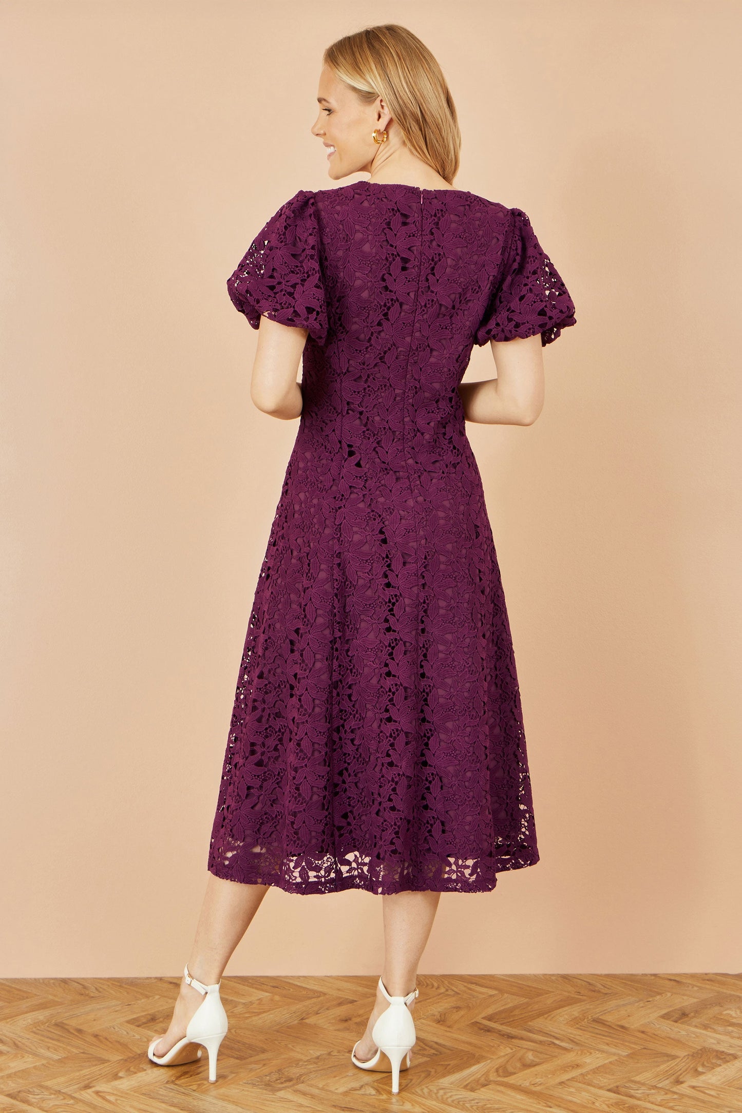 Woman wearing a purple lace dress against a beige wall.