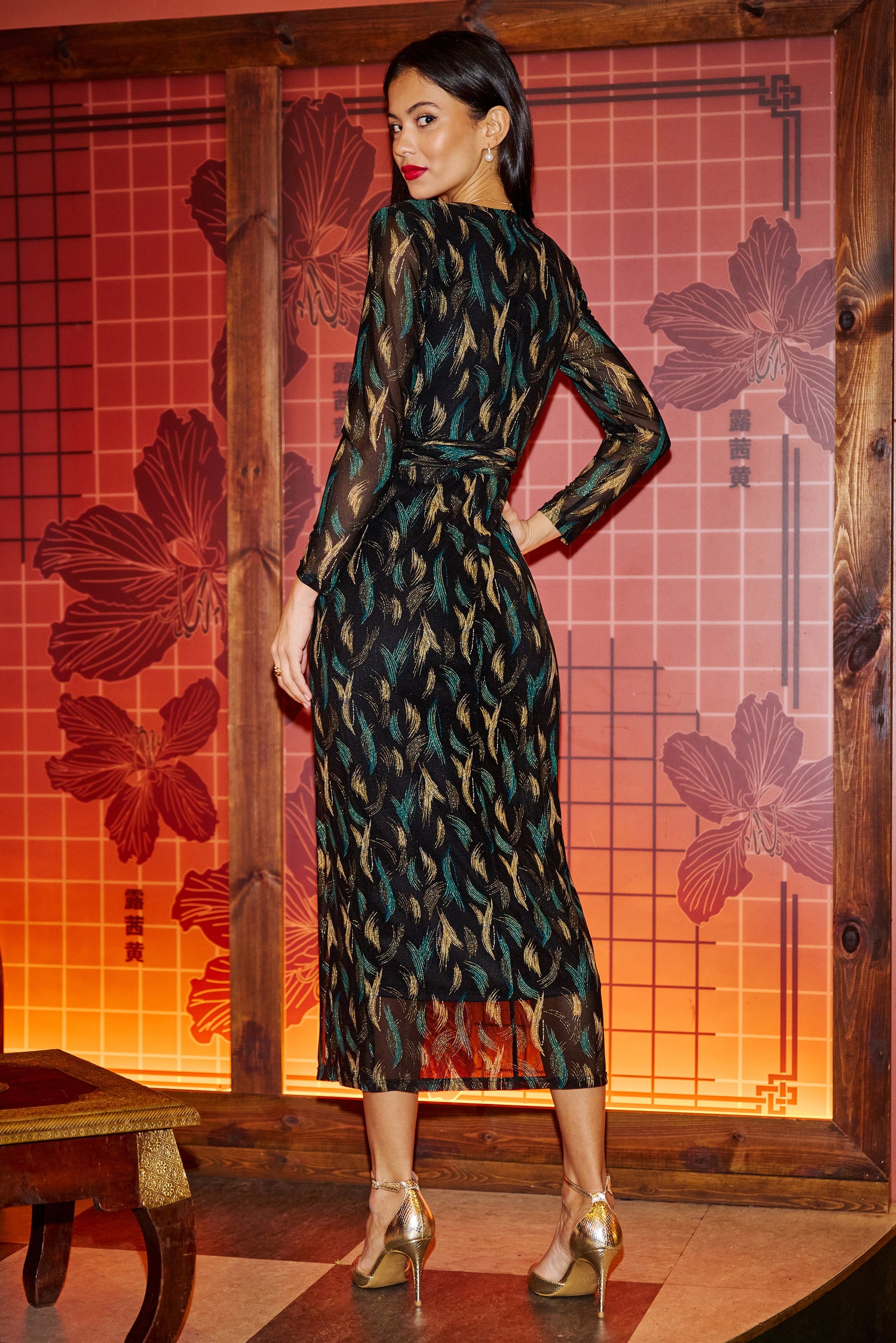 Woman in a patterned dress standing in front of a decorative wall with floral designs.