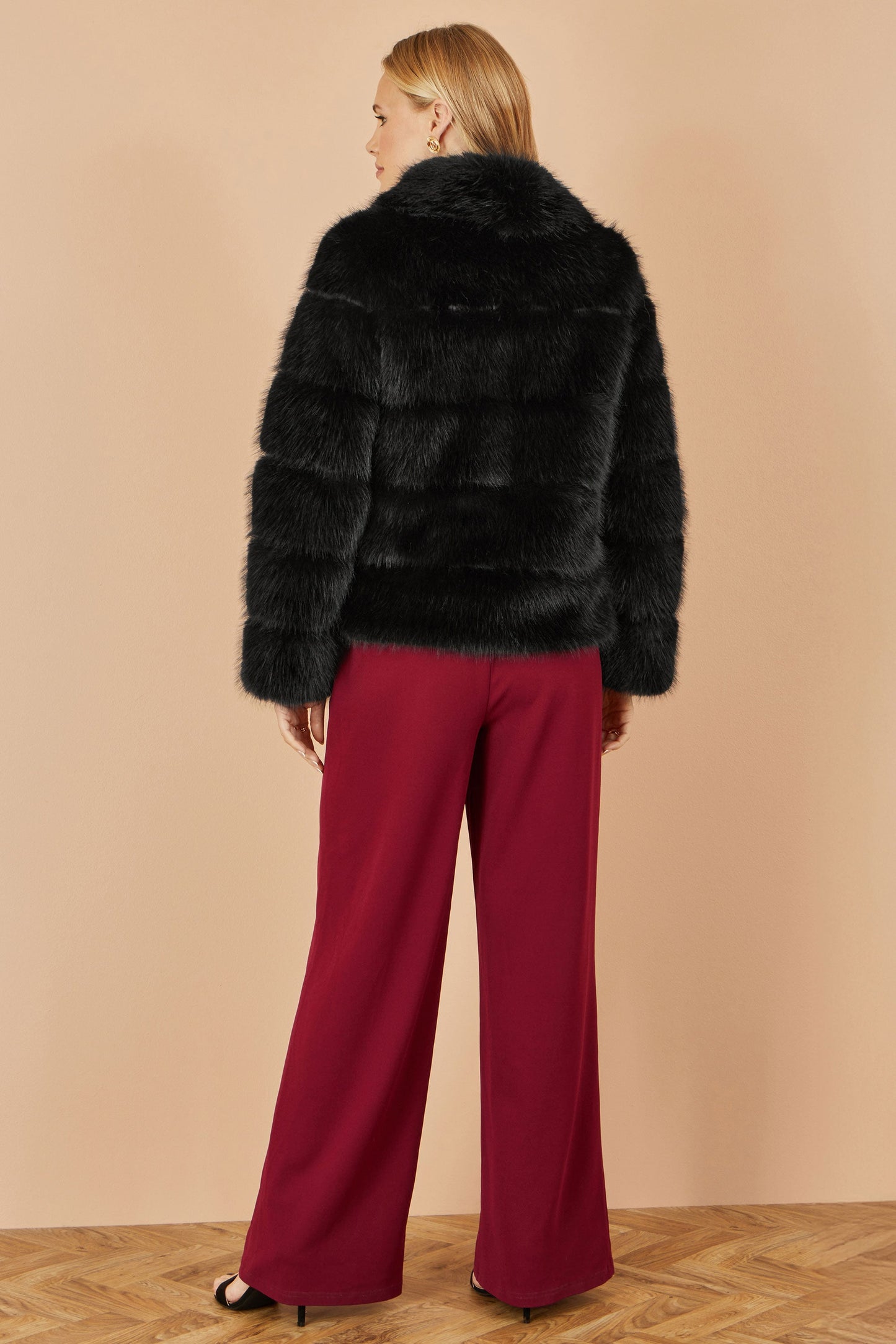 Person wearing a black fur coat and red pants on a beige background
