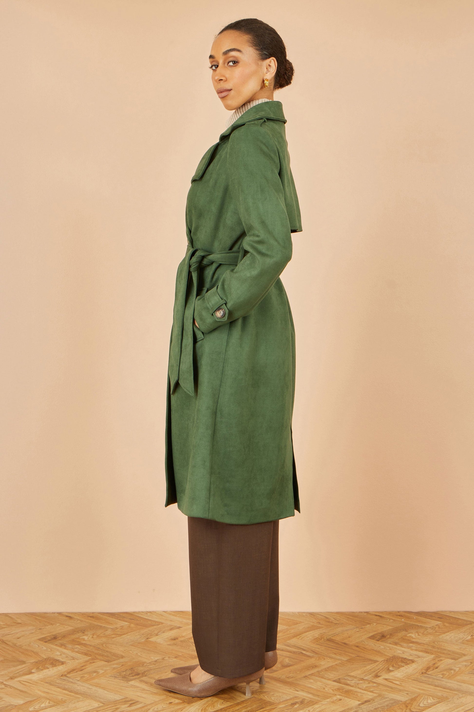 Woman wearing a green coat standing on a wooden floor with a beige wall background