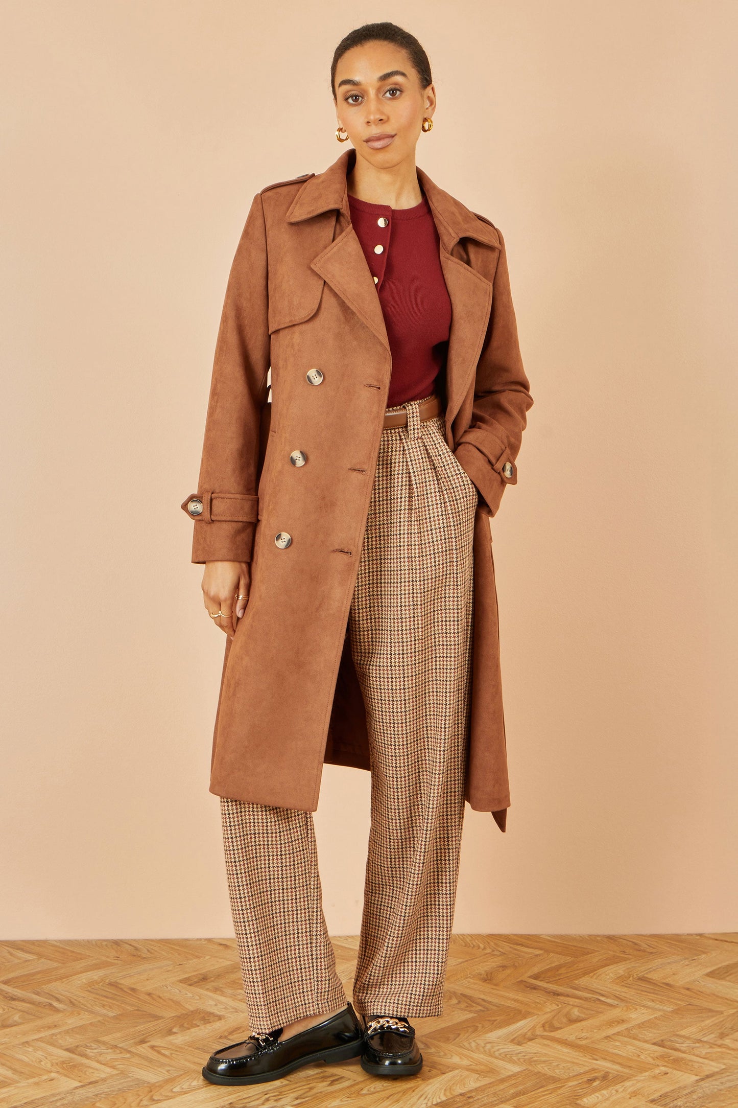 Woman wearing a brown coat, red top, and plaid pants on a beige background
