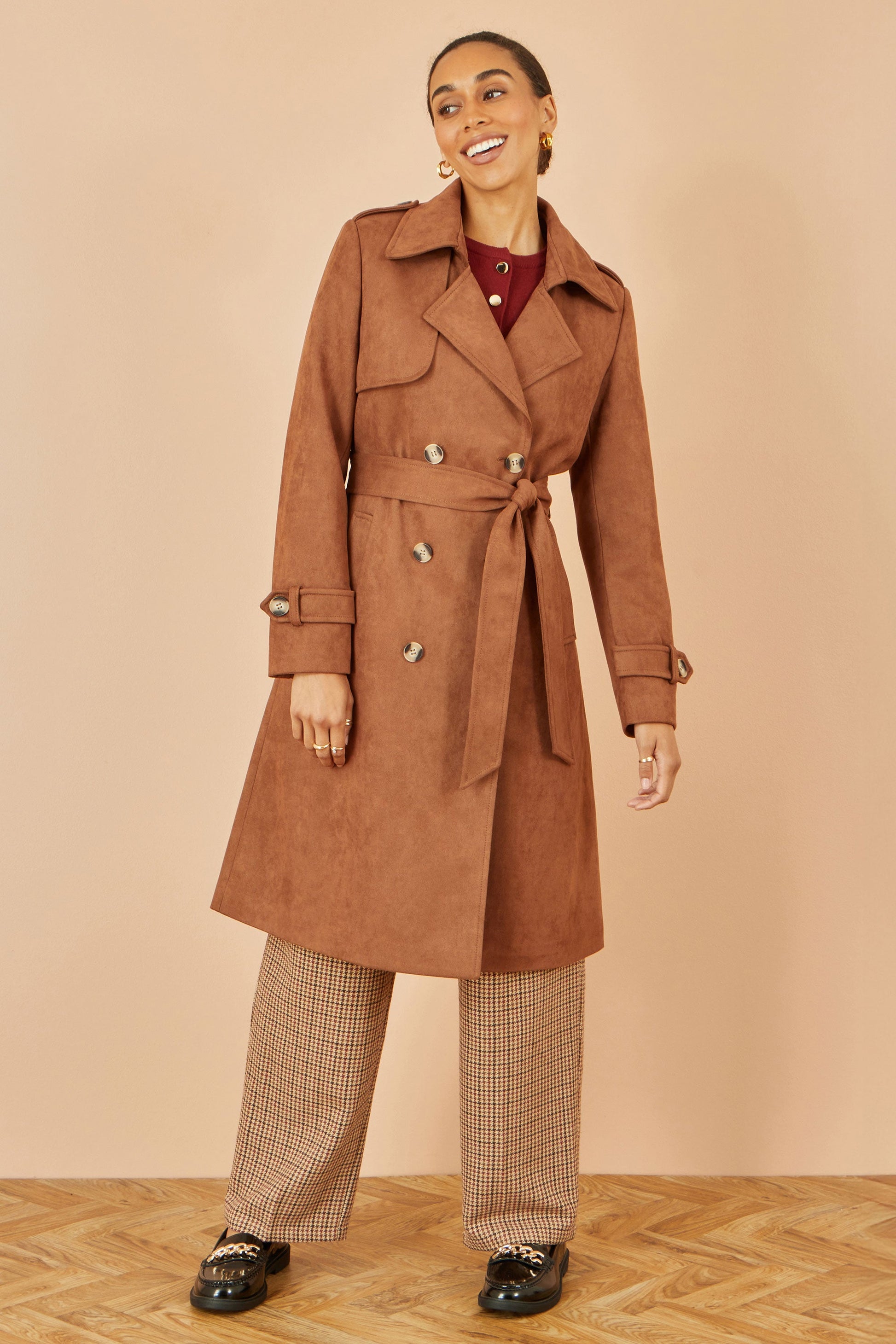 Woman wearing a brown trench coat with plaid pants against a beige background