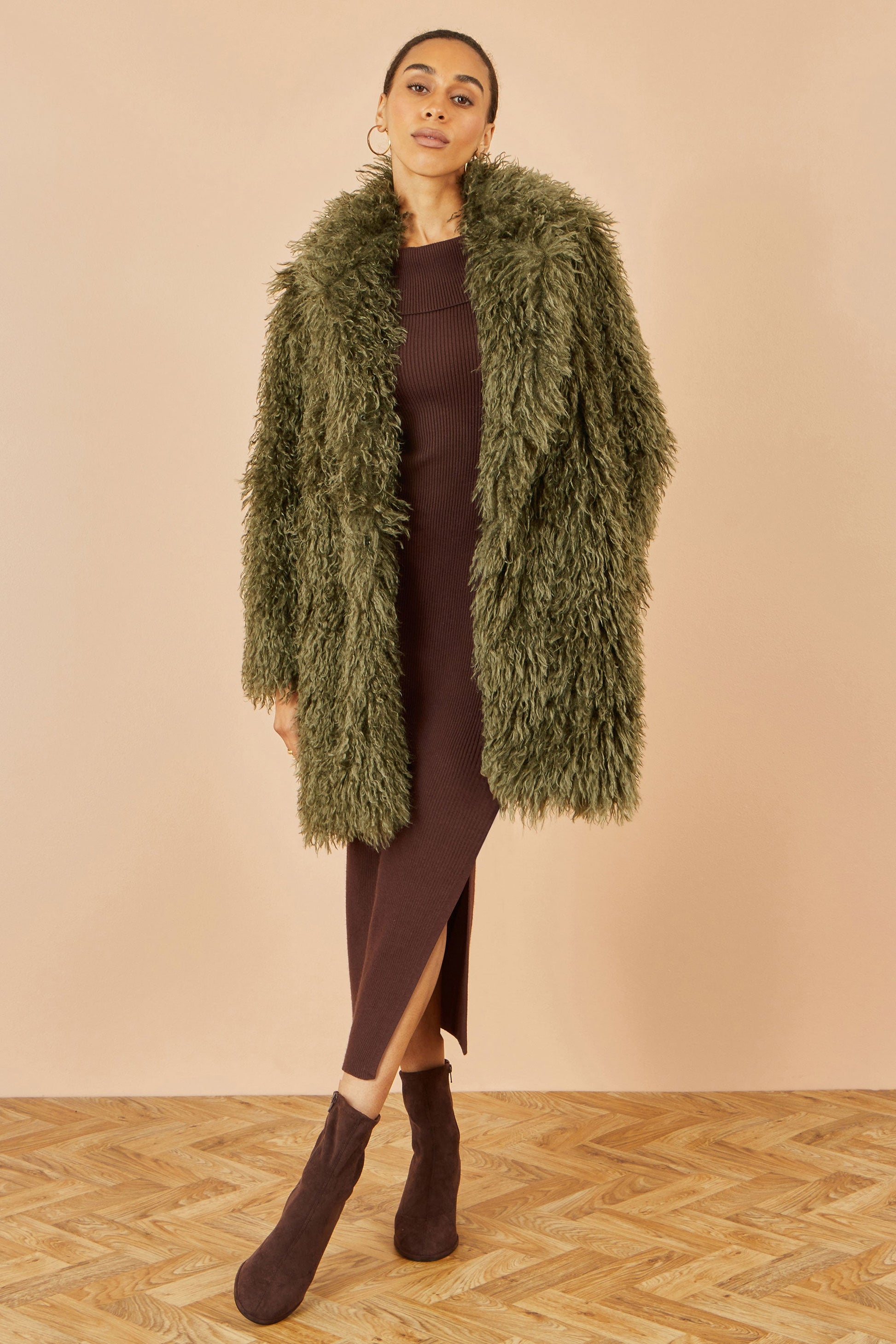 Woman wearing a long green fur coat over a brown dress on a beige background