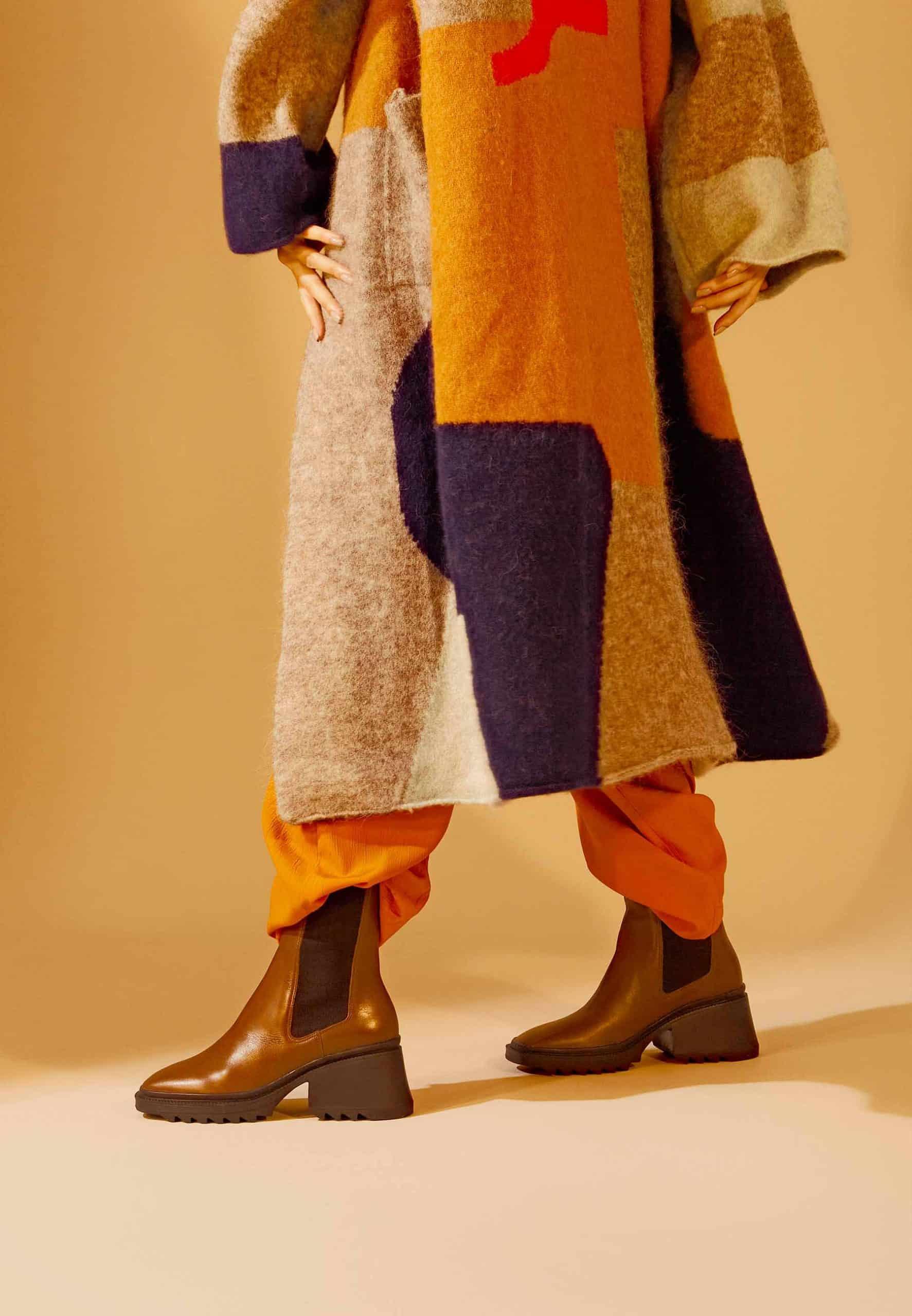 Person wearing a colorful patchwork coat and brown boots on a beige background