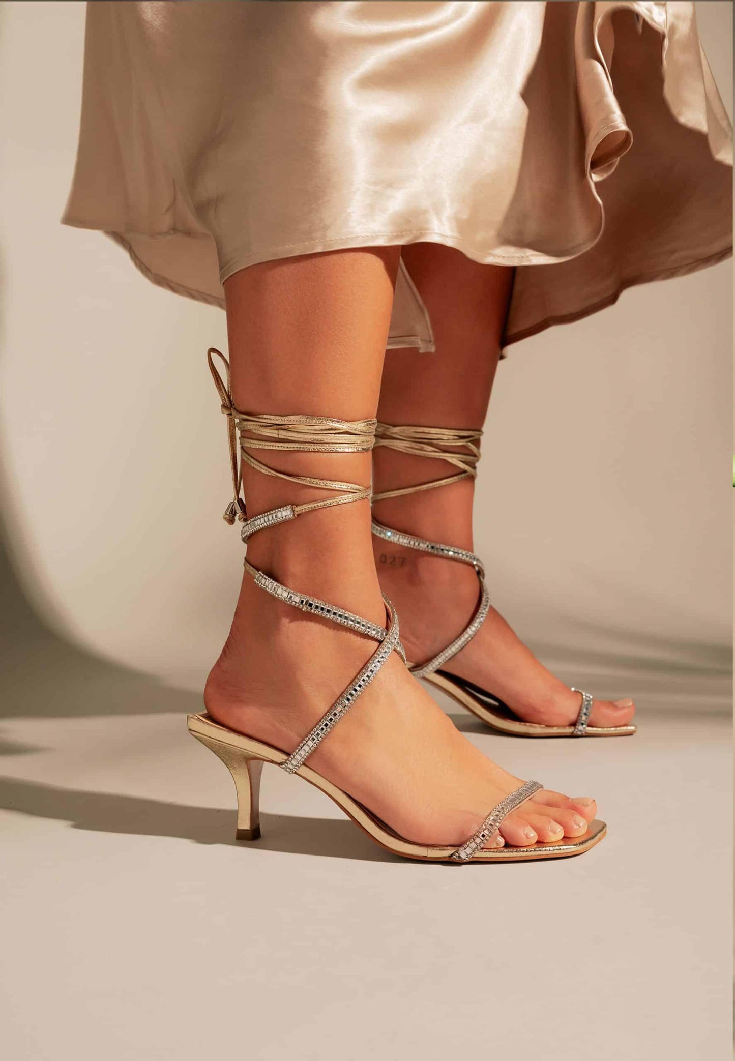 Beige high-heeled sandals with strappy design worn by a person in a light-colored dress.