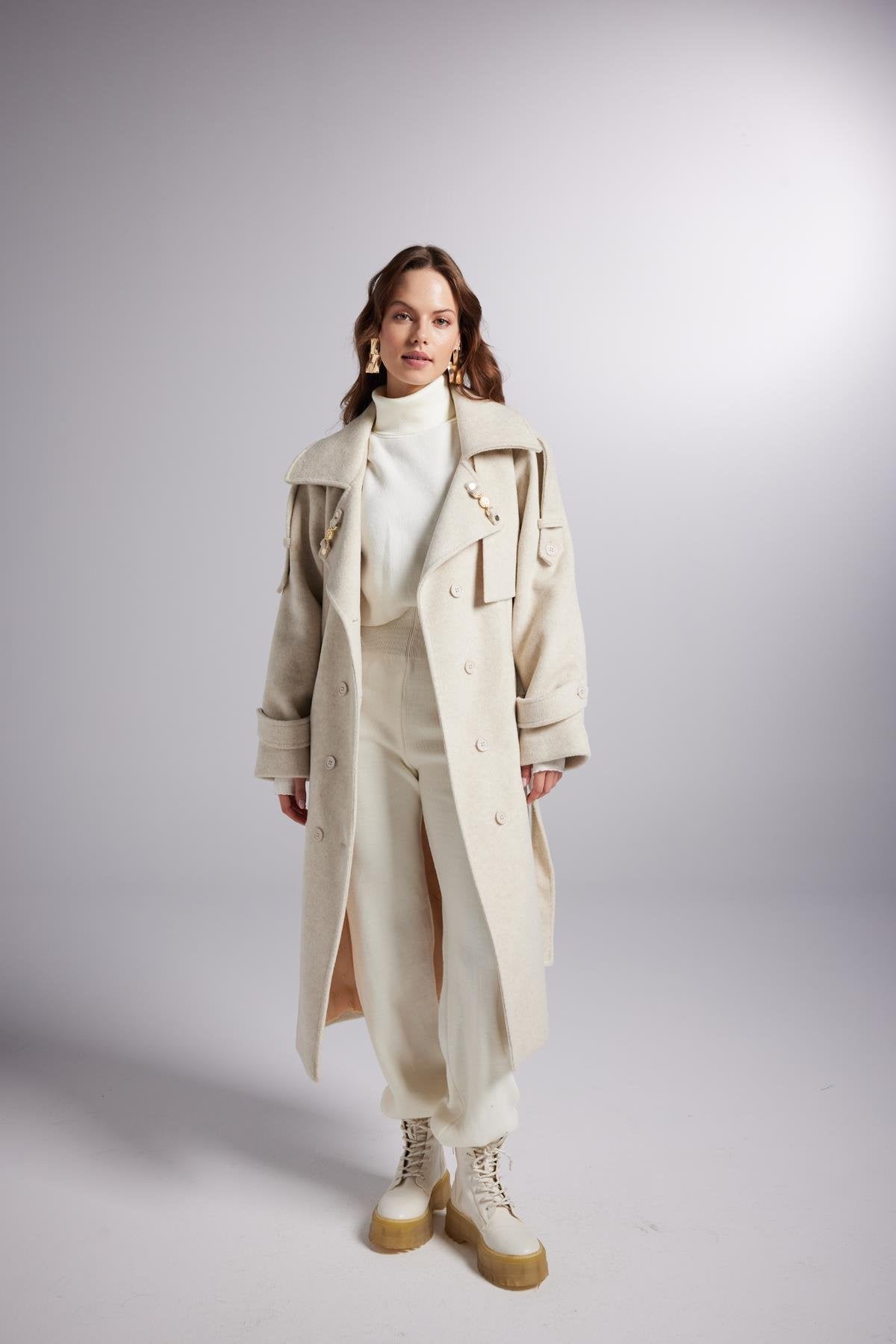 Woman wearing a long beige coat over a white outfit on a gray background