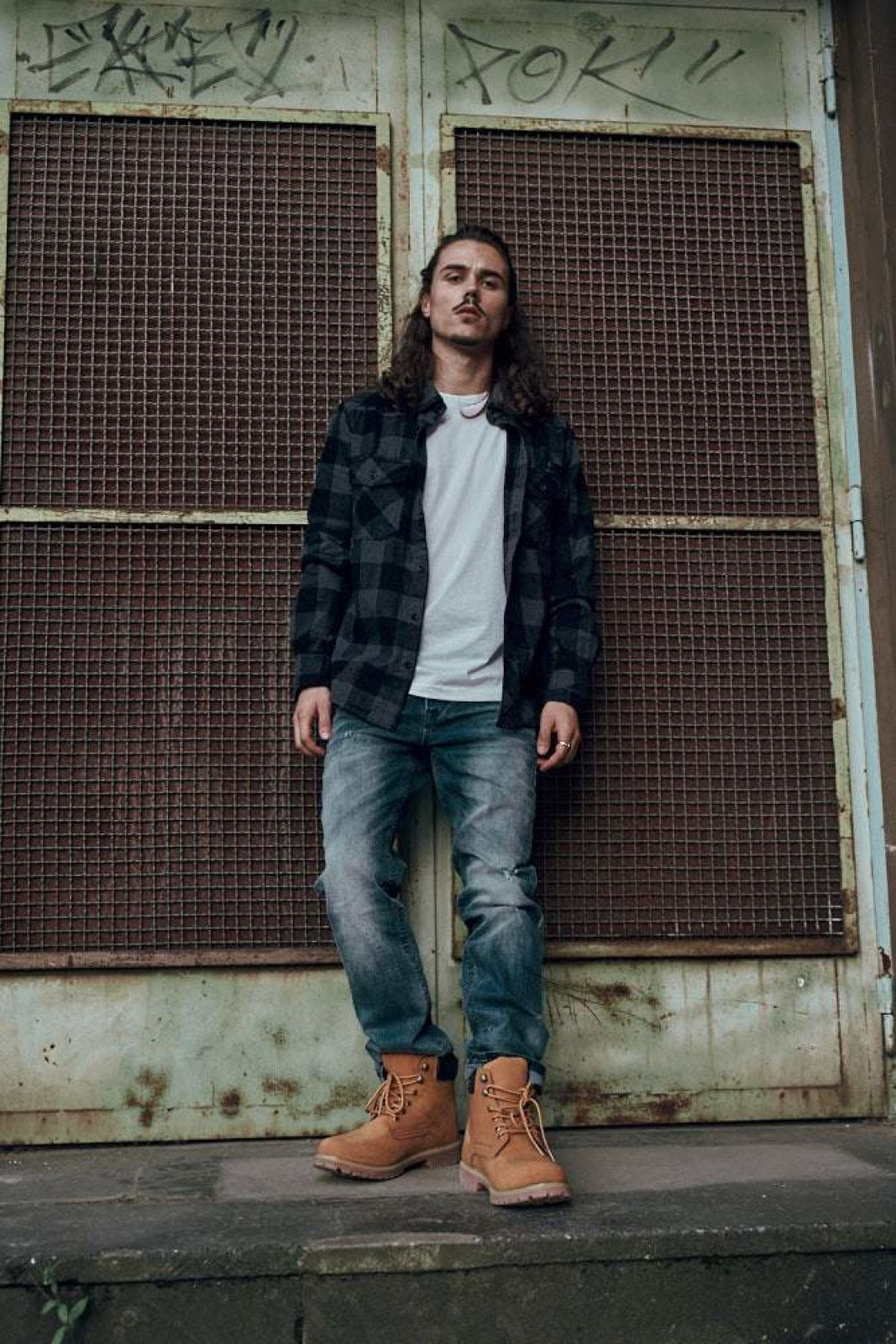 Person wearing a plaid shirt, white t-shirt, jeans, and brown boots standing in front of a metal door with graffiti.