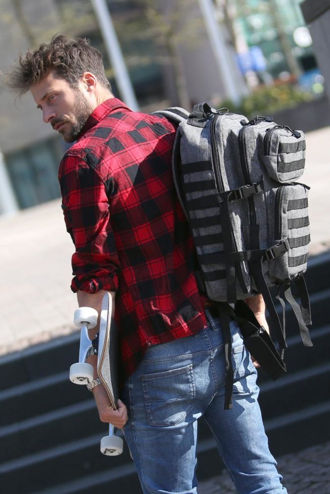 Man wearing a red plaid shirt and blue jeans with multiple backpacks on his back, holding a skateboard.