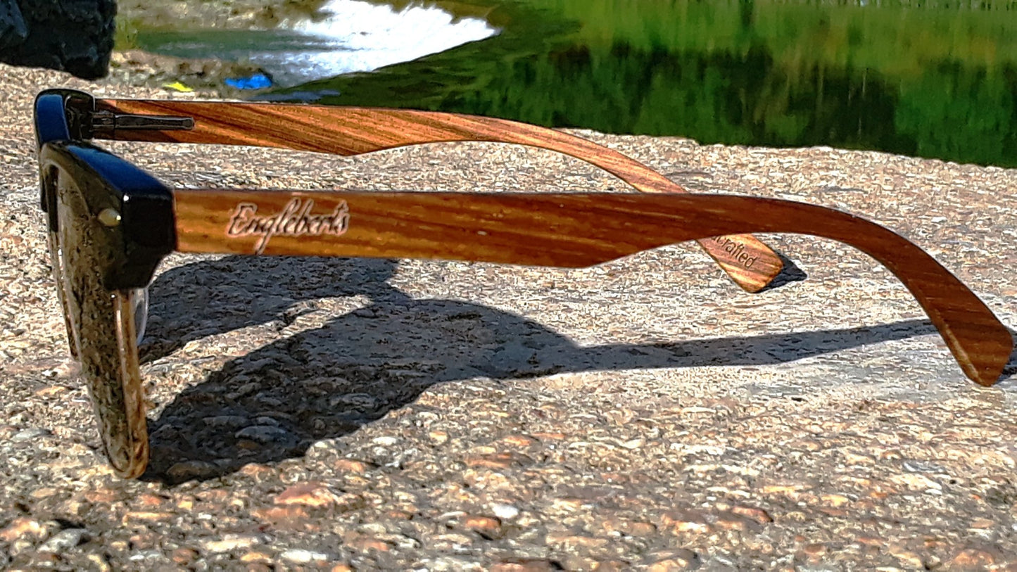 Wooden sunglasses with branding on a stone surface near water.